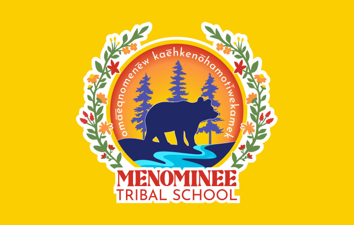 Logo for Menominee Tribal School featuring a bear silhouette standing by a river with pine trees in the background, surrounded by a circular border with floral decorations. The text “Menominee Tribal School” appears below, and Menominee language text is written around the top of the circle.