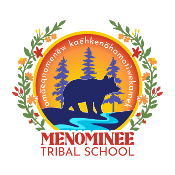 A circular logo for Menominee Tribal School. The center features a silhouette of a bear walking beside a blue river, with pine trees and an orange sunset in the background. Surrounding the circle are floral branches with red, yellow, and white flowers. The Menominee language phrase is written in a curved line above the bear. Below the circle, the text reads “MENOMINEE TRIBAL SCHOOL” in bold red and white letters.