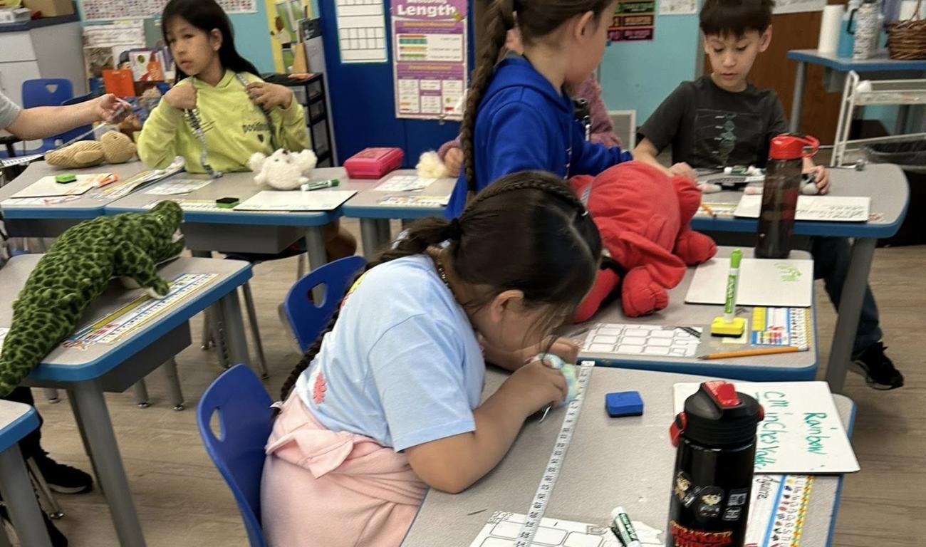 Children in a classroom use measuring tapes and stuffed animals at their desks, working on a hands-on activity.
