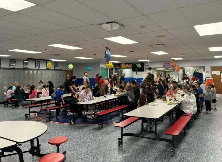 A busy school cafeteria with students and adults seated at long tables, eating and talking. Colorful fish decorations hang from the ceiling, and lockers line the back wall. Some people are standing and interacting, while others are seated and enjoying their meals.