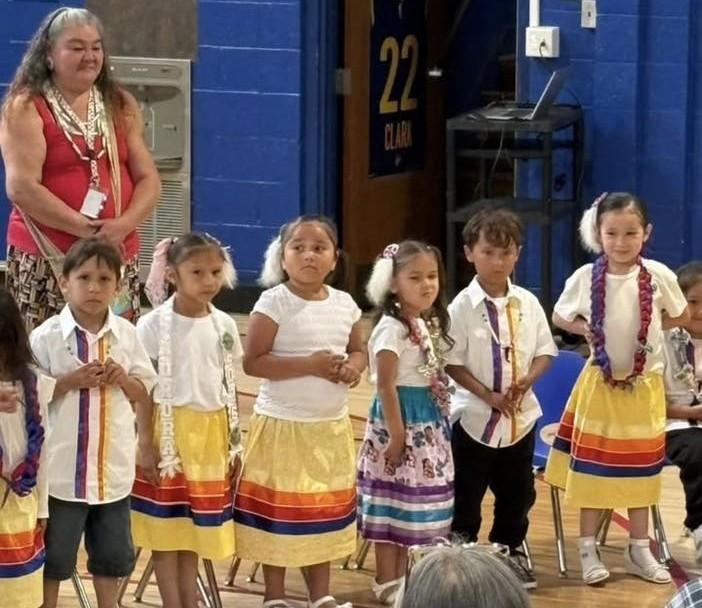 Young students at Menominee Tribal School dressed in traditional clothing, standing in a row during a cultural event indoors, with an adult behind them.
