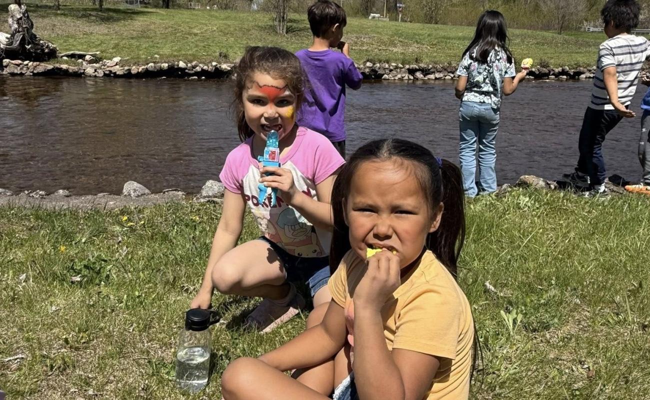 Students at Menominee Tribal School participating in outdoor activities near a stream, with two children in the foreground eating snacks and other students standing by the water.