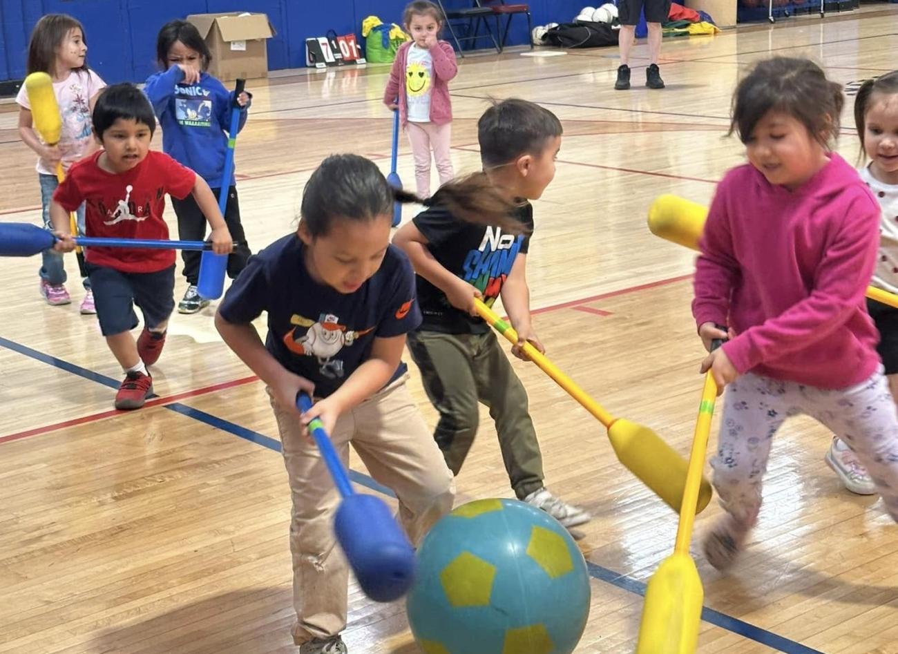 Students at Menominee Tribal School participating in physical activity indoors, playing with foam bats and a large ball in a gymnasium.